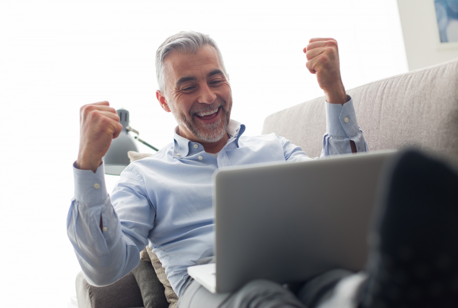 Cheerful businessman lying on the couch at home and using a laptop, he is joyful and raising his fists